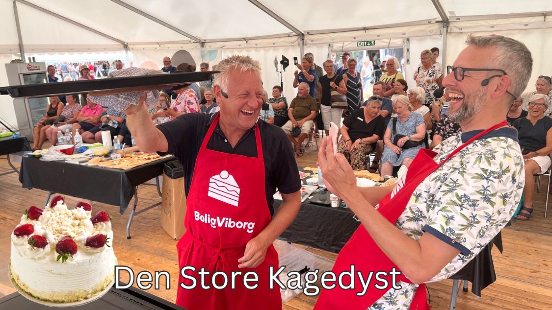 På billedet ses to mænd, der deltager i "Teambuilding København: Den Store Kagedyst". De er begge klædt i røde forklæder og ser ud til at have det sjovt. Den ene mand holder en bakke med kager højt oppe, mens den anden klapper og smiler bredt. Bag dem er der et publikum, som også nyder arrangementet. Der står en smuk kage på bordet foran dem, pyntet med flødeskum og jordbær. Billedteksten "Den Store Kagedyst"