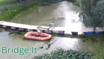 To personer bygger en flydende bro over en sø ved hjælp af planker og tønder. En deltager står i en orange gummibåd, mens den anden arbejder på broen. Omgivet af grøn natur. Billedetekst: "Bridge It